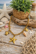 Collier Procida en acier inoxydable doré avec charms marins, étoile de mer, tortue, coquillages et détail rose sur décor plage.