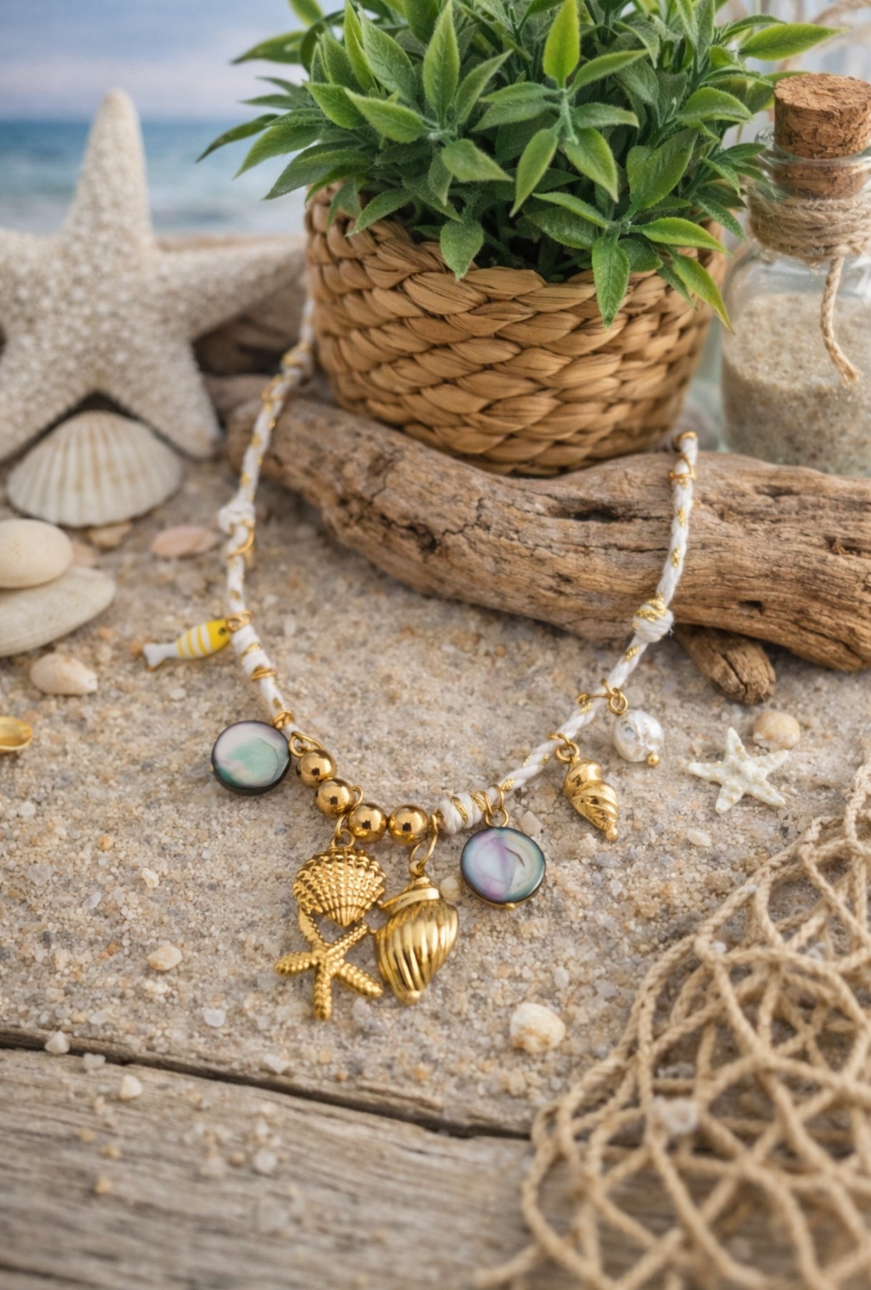 Collier Ischia en acier inoxydable doré avec cordon blanc, charms coquillages, étoile de mer, perles, poisson et détails nacrés sur décor plage.
