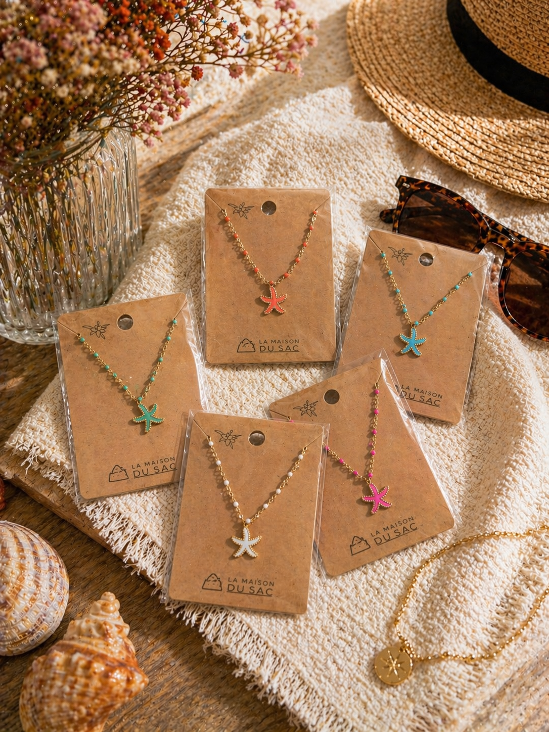Colliers dorés avec pendentif étoile de mer colorée, présentés sur cartes La Maison du Sac dans une ambiance estivale méditerranéenne avec coquillages, chapeau de paille et lunettes de soleil.