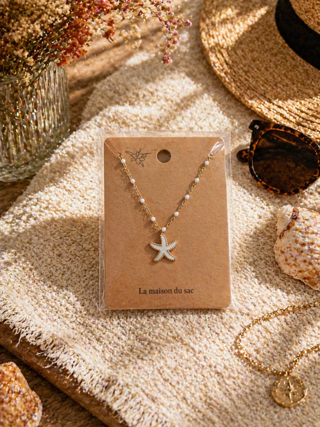 Colliers dorés avec pendentif étoile de mer colorée, présentés sur cartes La Maison du Sac dans une ambiance estivale méditerranéenne avec coquillages, chapeau de paille et lunettes de soleil.