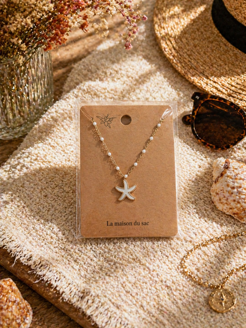 Colliers dorés avec pendentif étoile de mer colorée, présentés sur cartes La Maison du Sac dans une ambiance estivale méditerranéenne avec coquillages, chapeau de paille et lunettes de soleil.